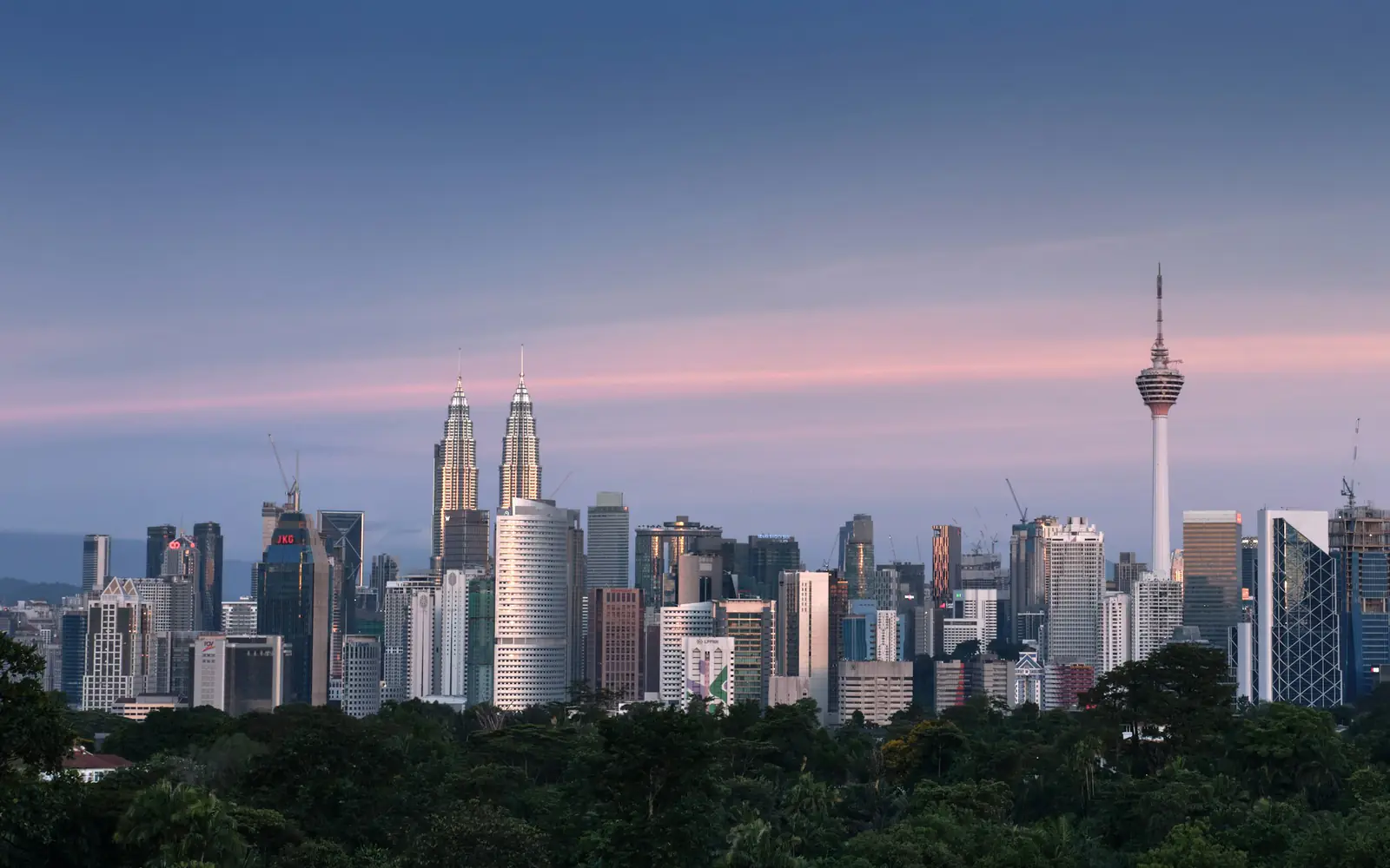 The Kuala Lumpur skyline at dusk, including the Petronas Towers and KL Tower, above a band of green trees.
