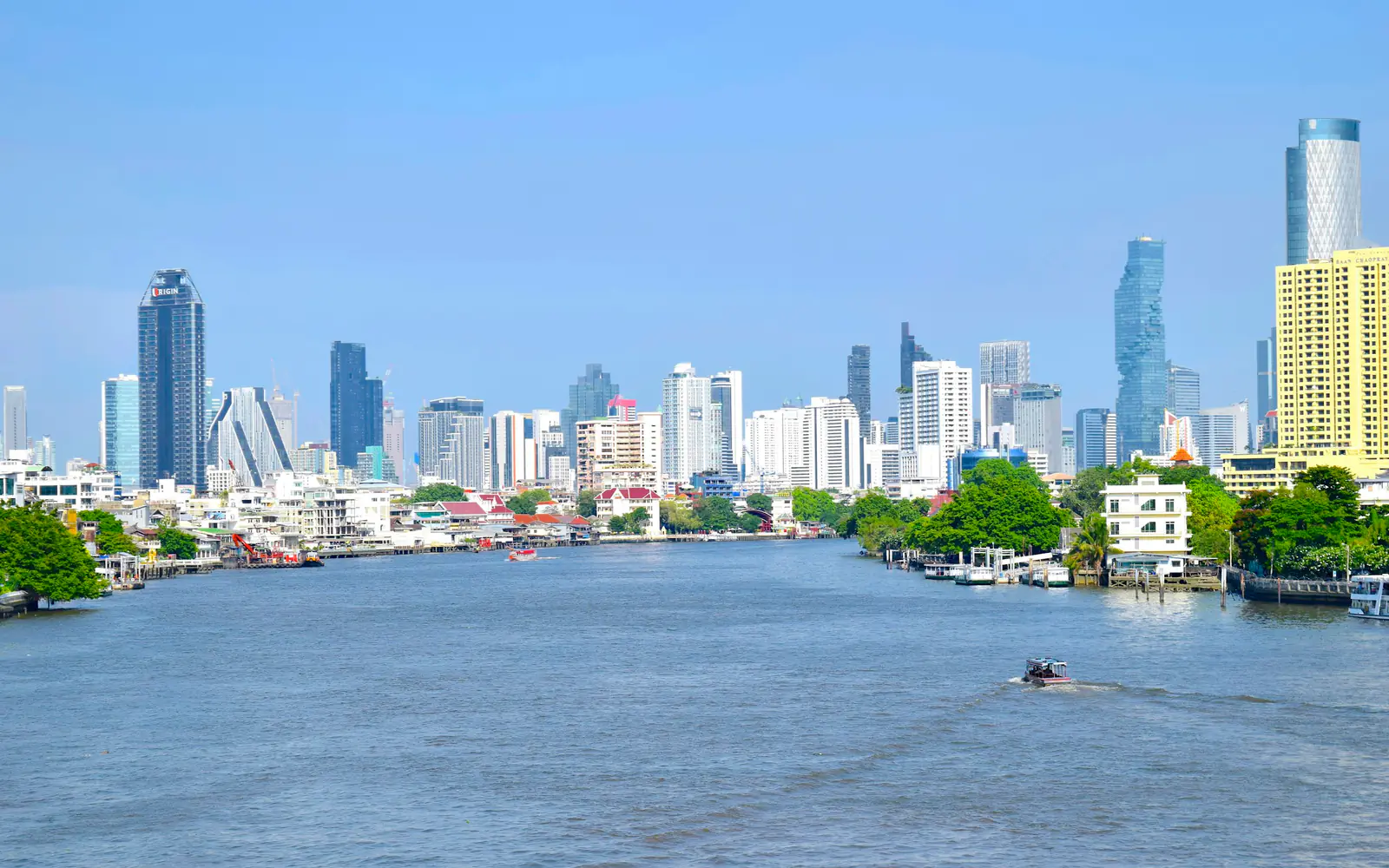 The Chao Phraya River in Bangkok with riverside trees and the city's high-rise skyline under a blue sky.
