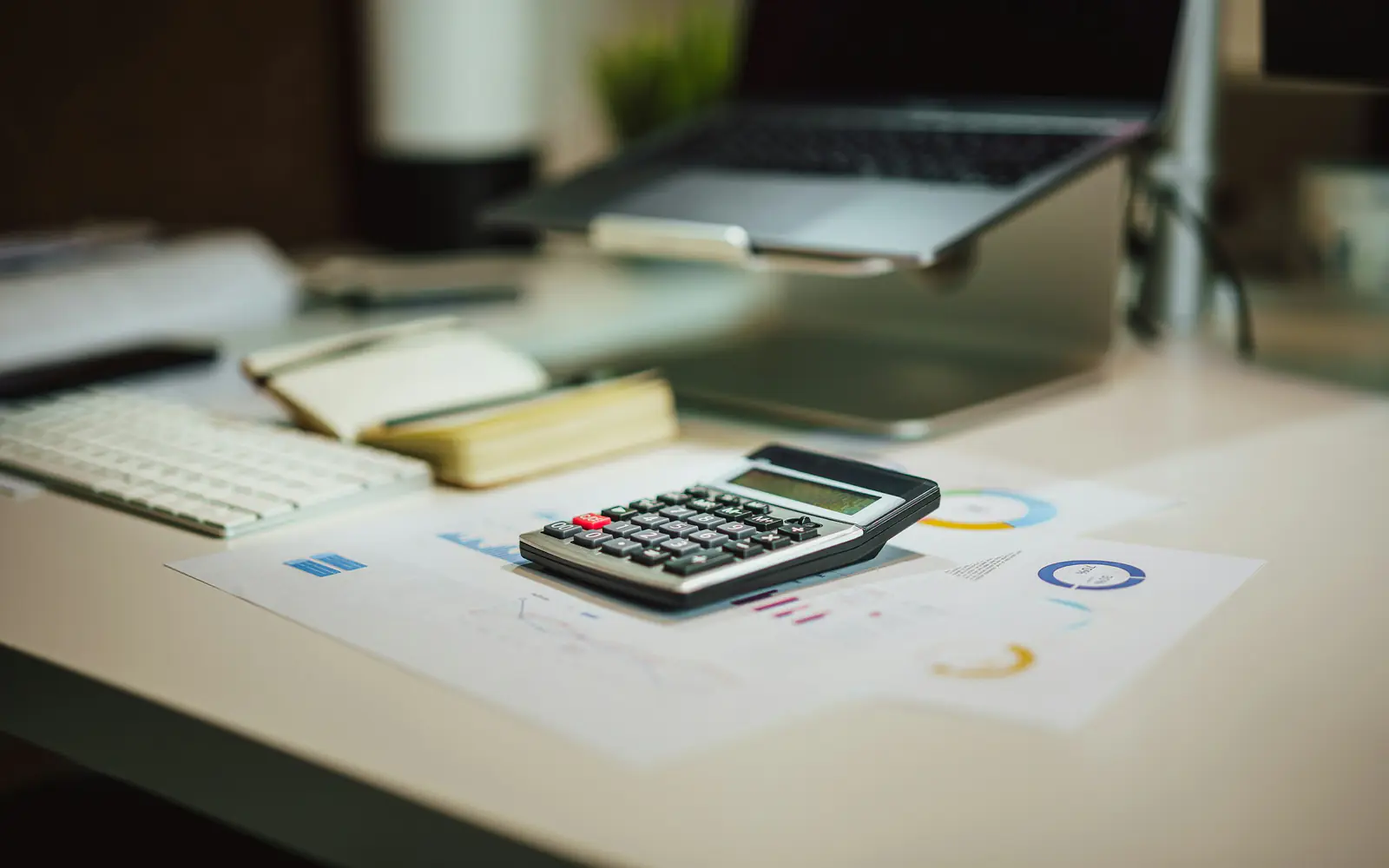 A calculator on printed charts beside a laptop and notebook on a desk.