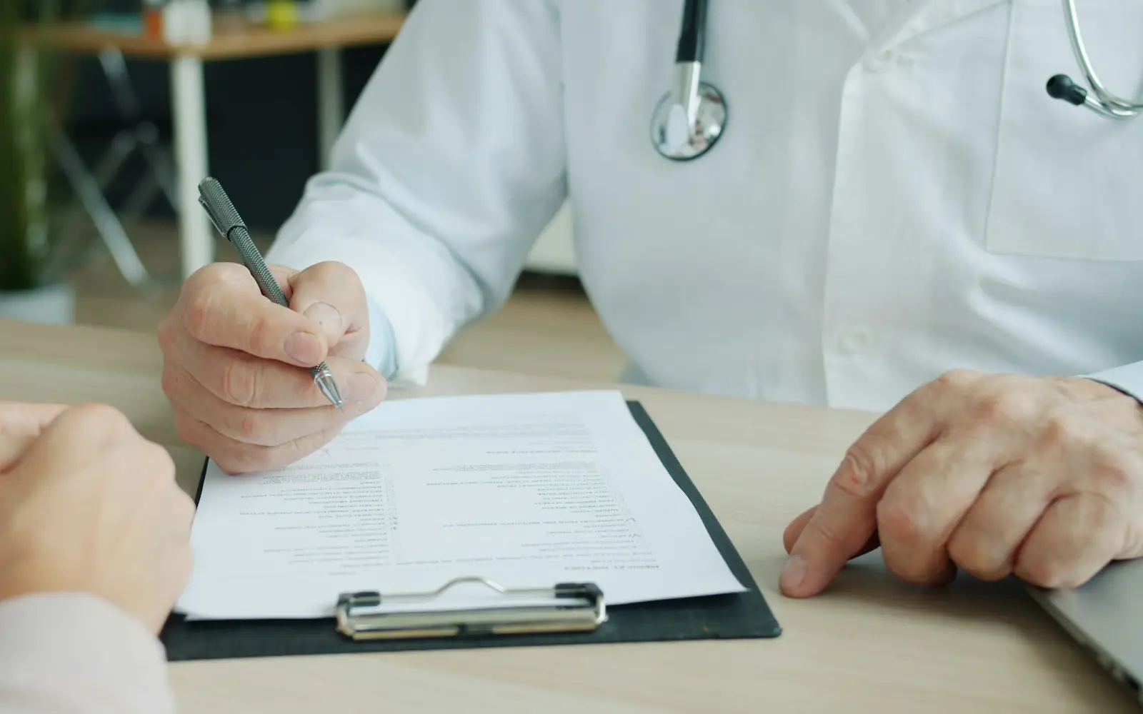 A doctor fills in a form on a clipboard during an appointment, with the patient's hands resting on the desk.