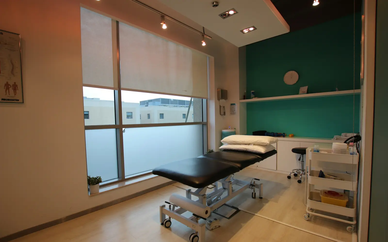 A physiotherapy room with a treatment table, an equipment trolley and a large window.