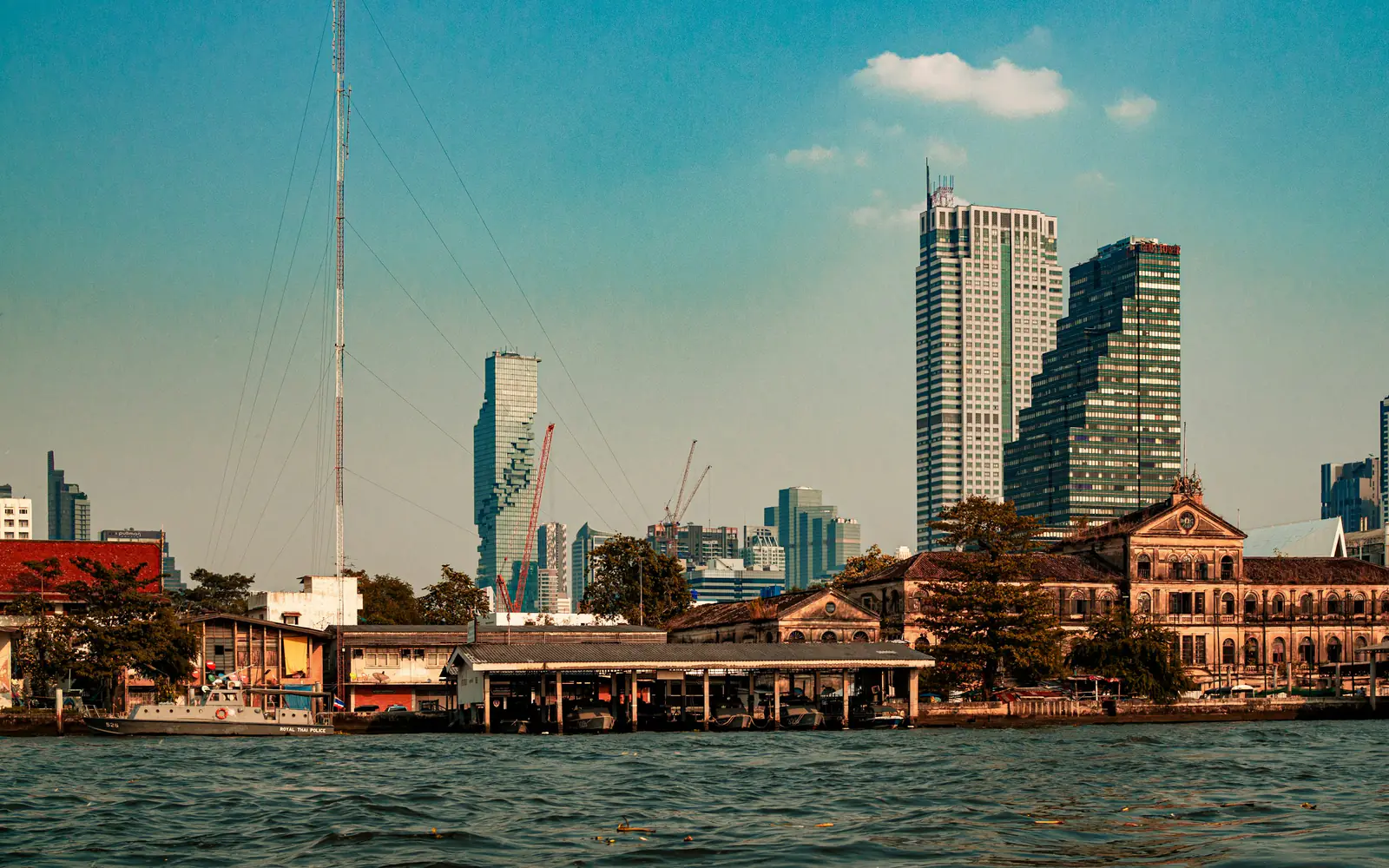 Riverside buildings and modern towers along the Chao Phraya River in Bangkok on a clear day.