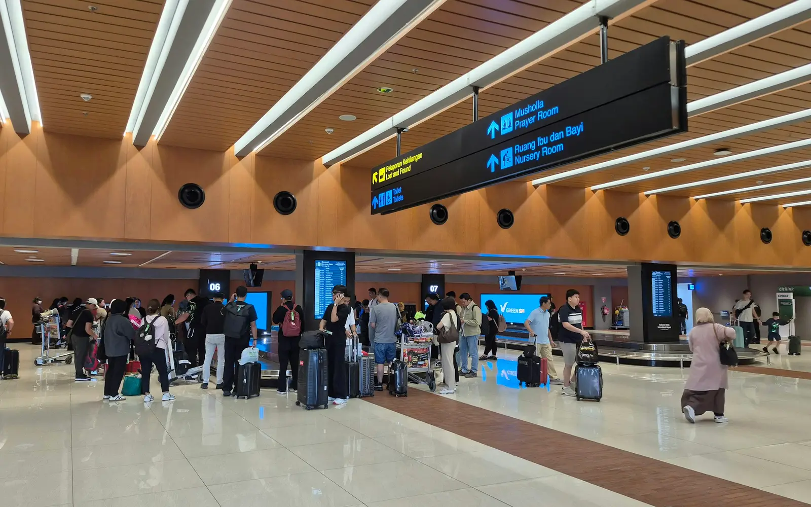 Travellers with suitcases and trolleys waiting at the baggage belts in an airport arrivals hall.