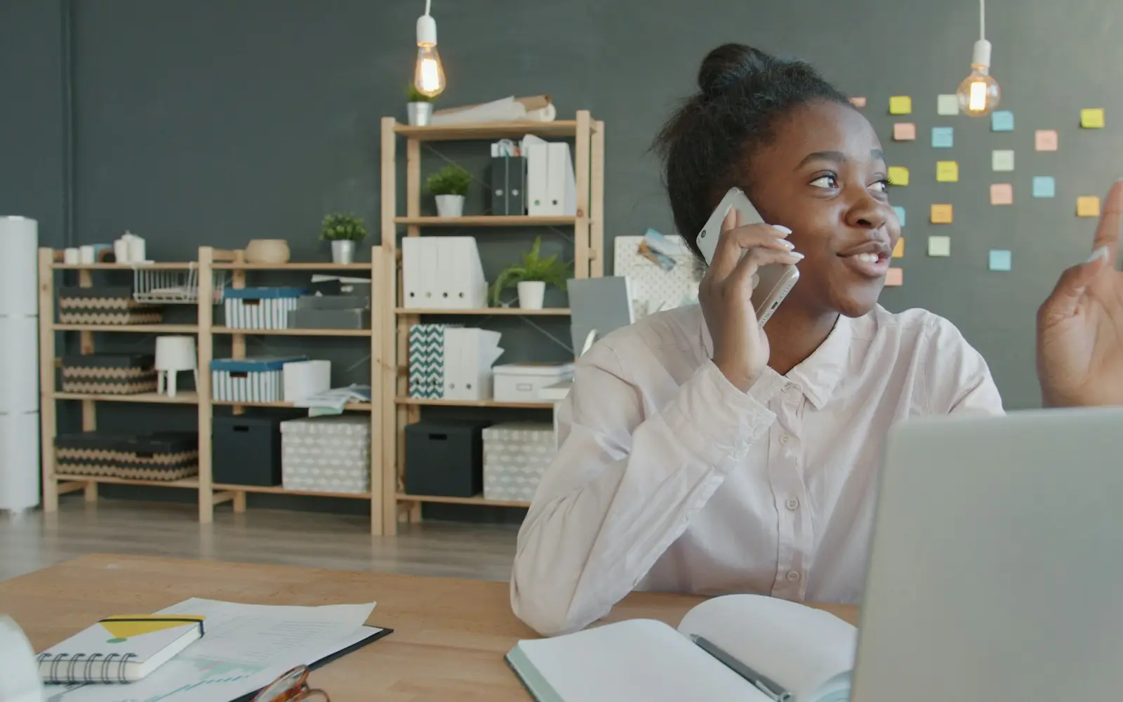 A young woman talks on the phone at a desk with a laptop and an open notebook.
