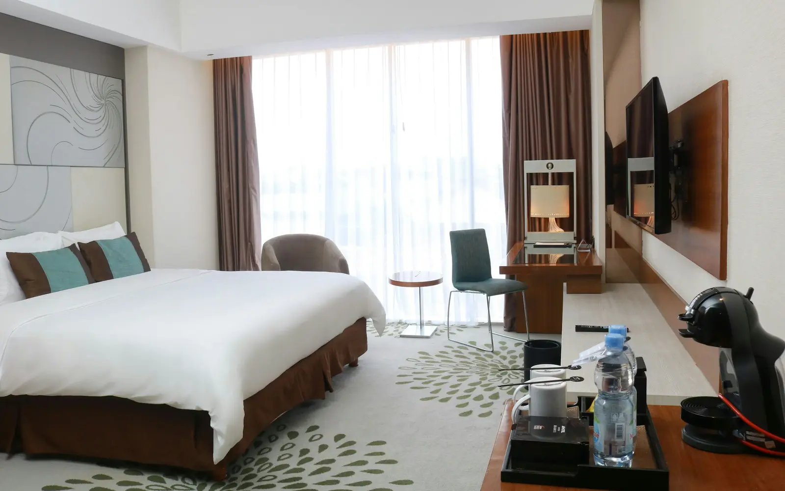 A bright hotel room with a double bed, an armchair, a desk and a large window with curtains.