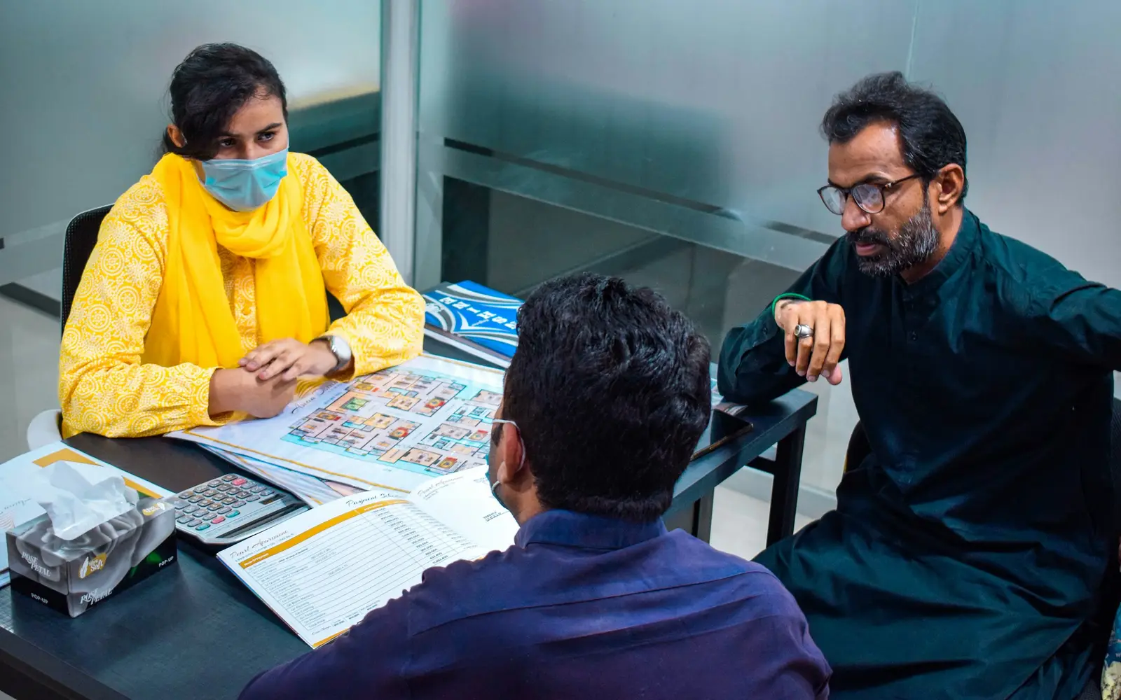 Three people talking across a desk covered with papers: a woman in a yellow scarf and two men.