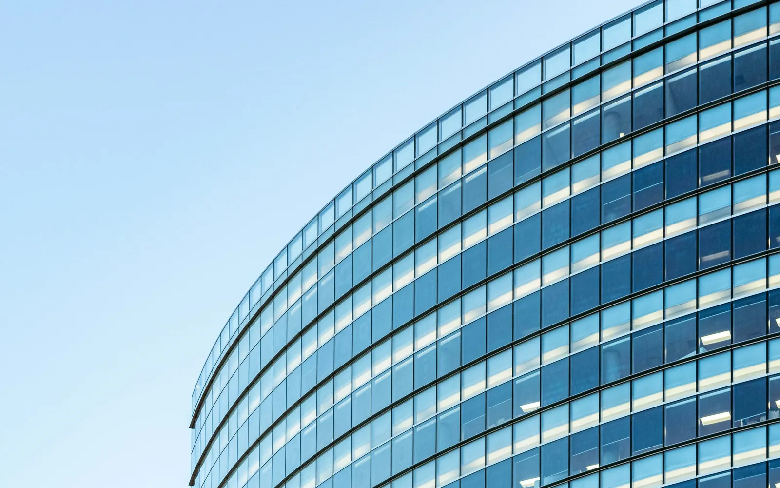 The curved glass facade of a modern office building against a clear blue sky.