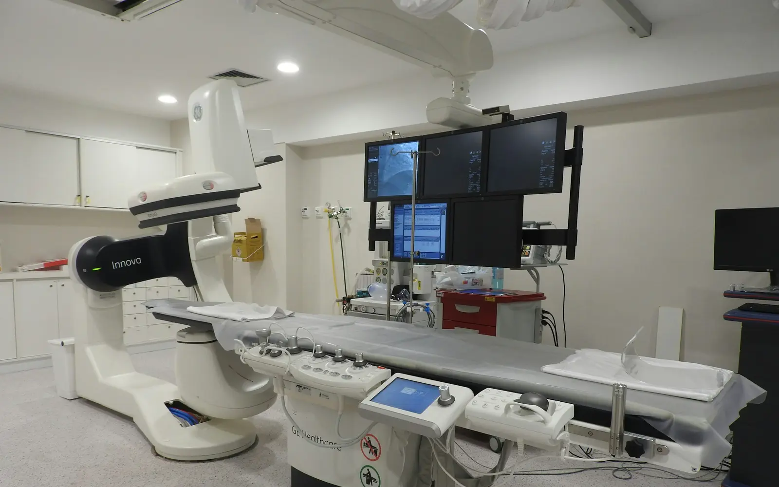 An empty cardiac catheterisation lab with an X-ray C-arm, a patient table and overhead monitors.