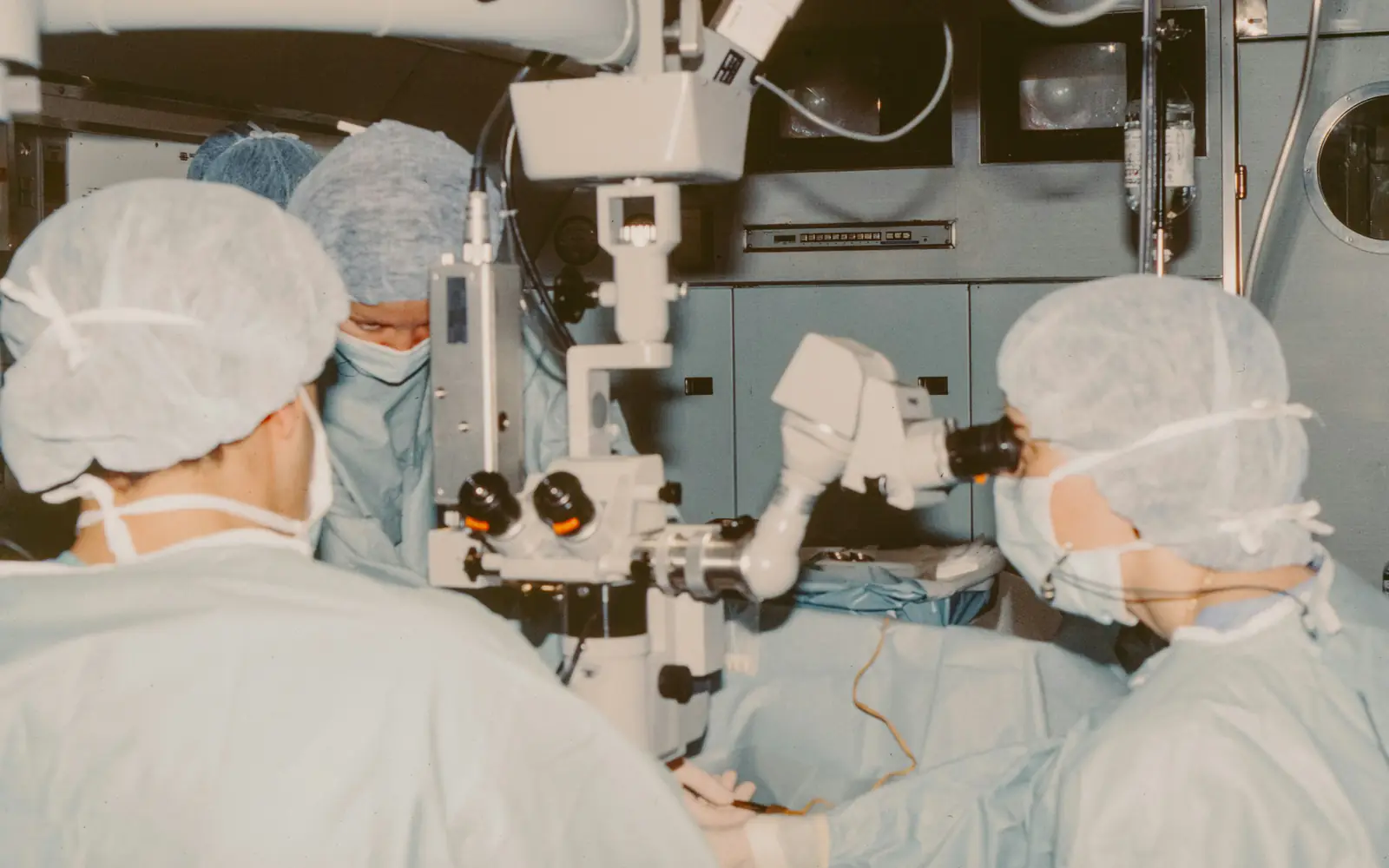 Surgeons in caps and masks operate using a large surgical microscope in an operating theatre.