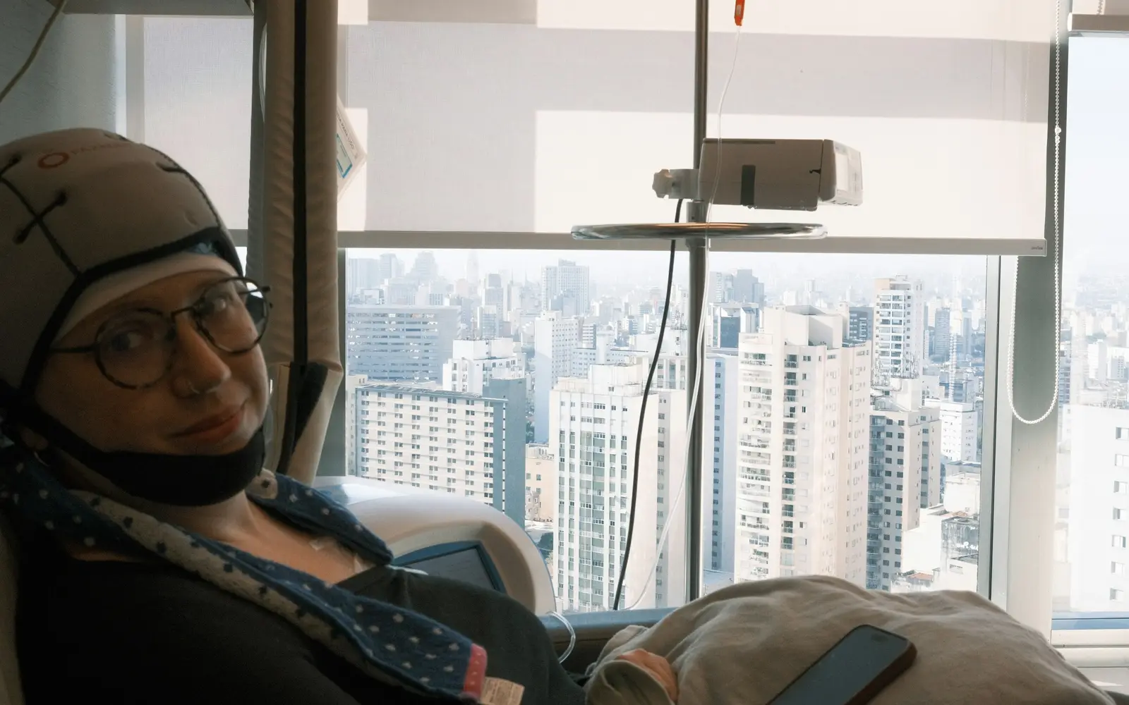 A woman wearing a scalp-cooling cap sits in a treatment chair by a window overlooking the city.