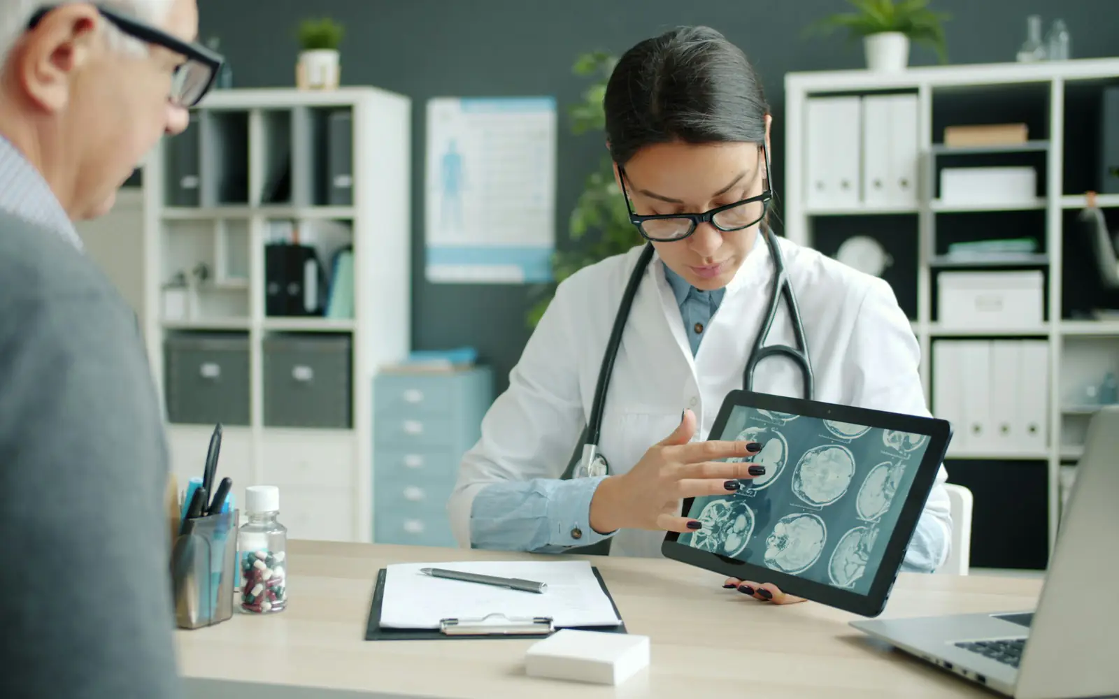 A doctor at her desk shows an older man brain scan images on a tablet.