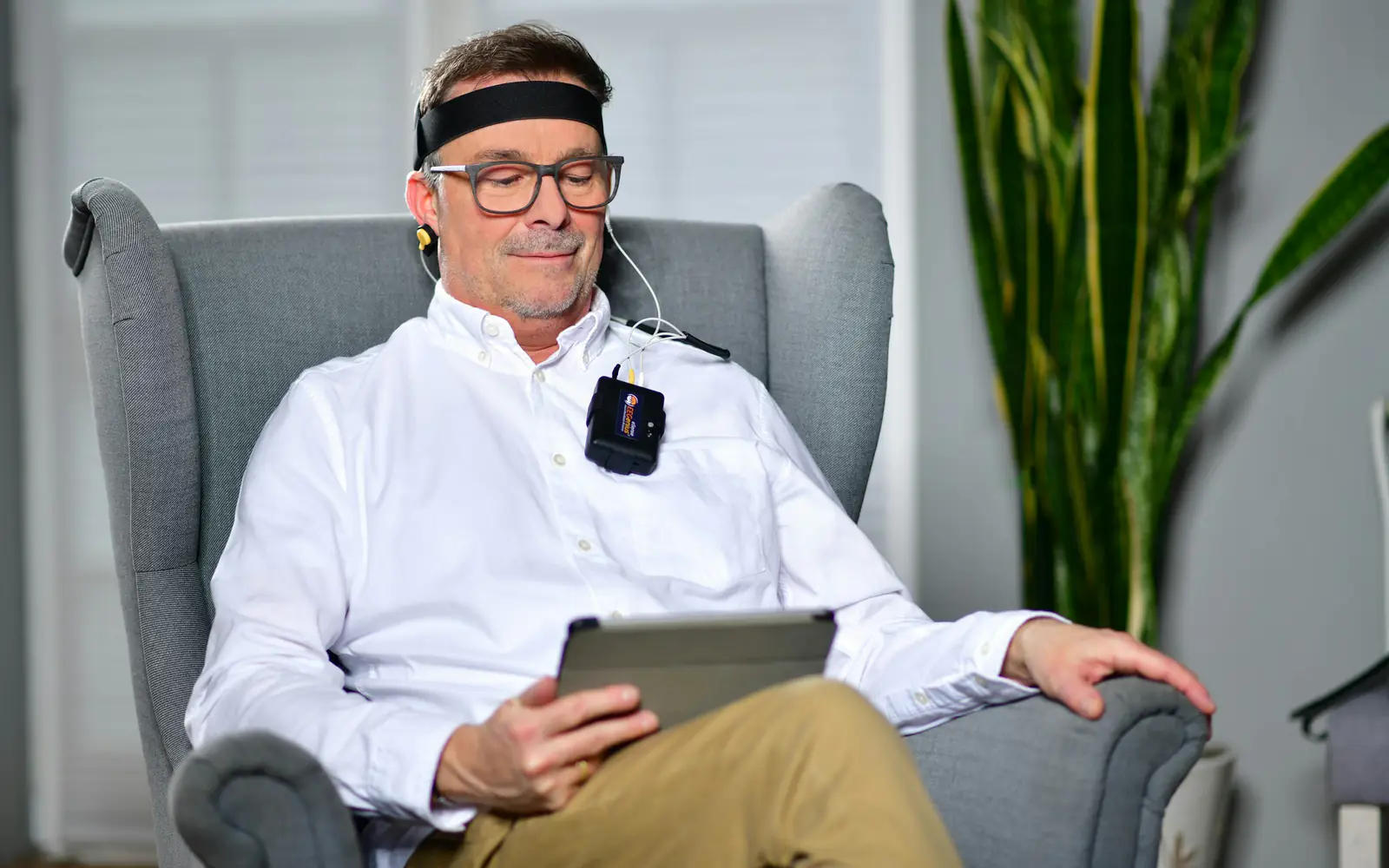 A man sits in an armchair wearing an EEG headband with electrodes while holding a tablet.
