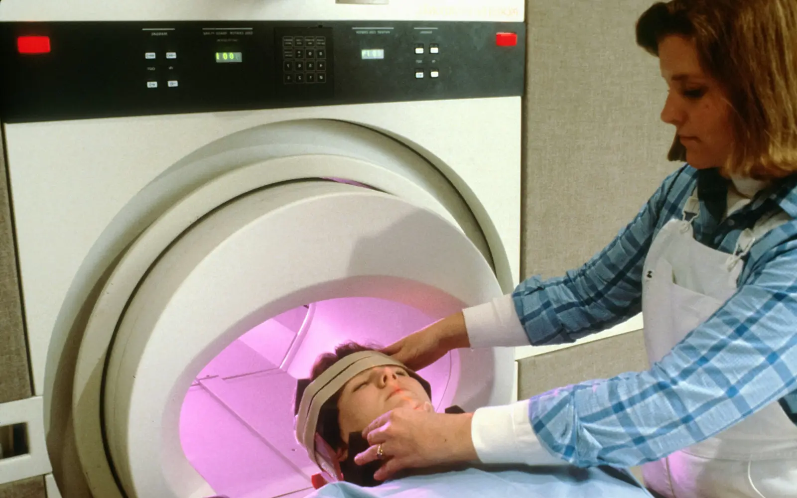 A radiation therapist positions a patient's head with a strap inside a radiotherapy machine.