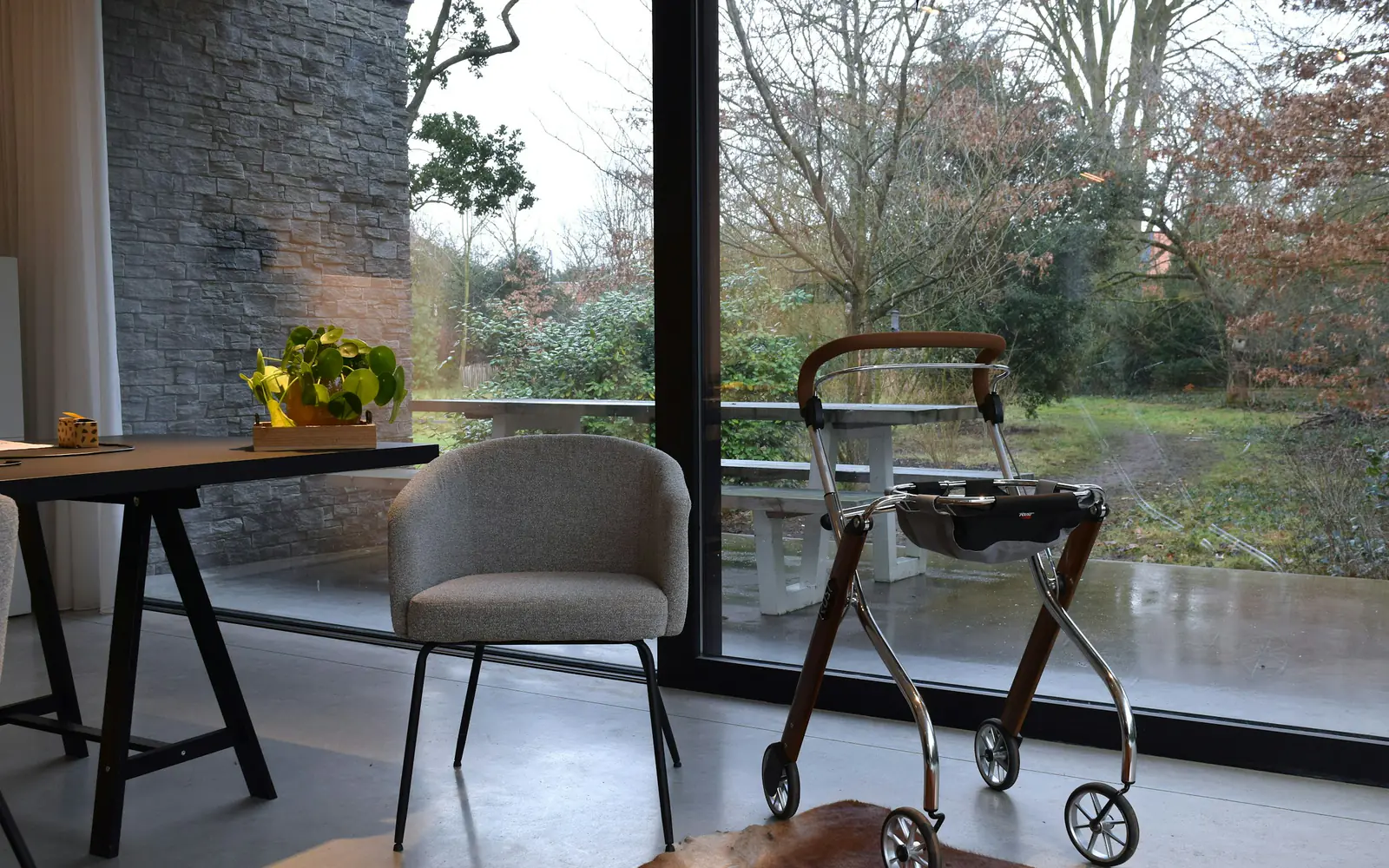 A wheeled walker stands beside a chair and table near a large window looking onto a garden.