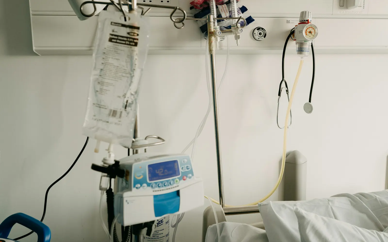 An infusion bag and pump on a drip stand beside a hospital bed, with a stethoscope hanging on the wall.