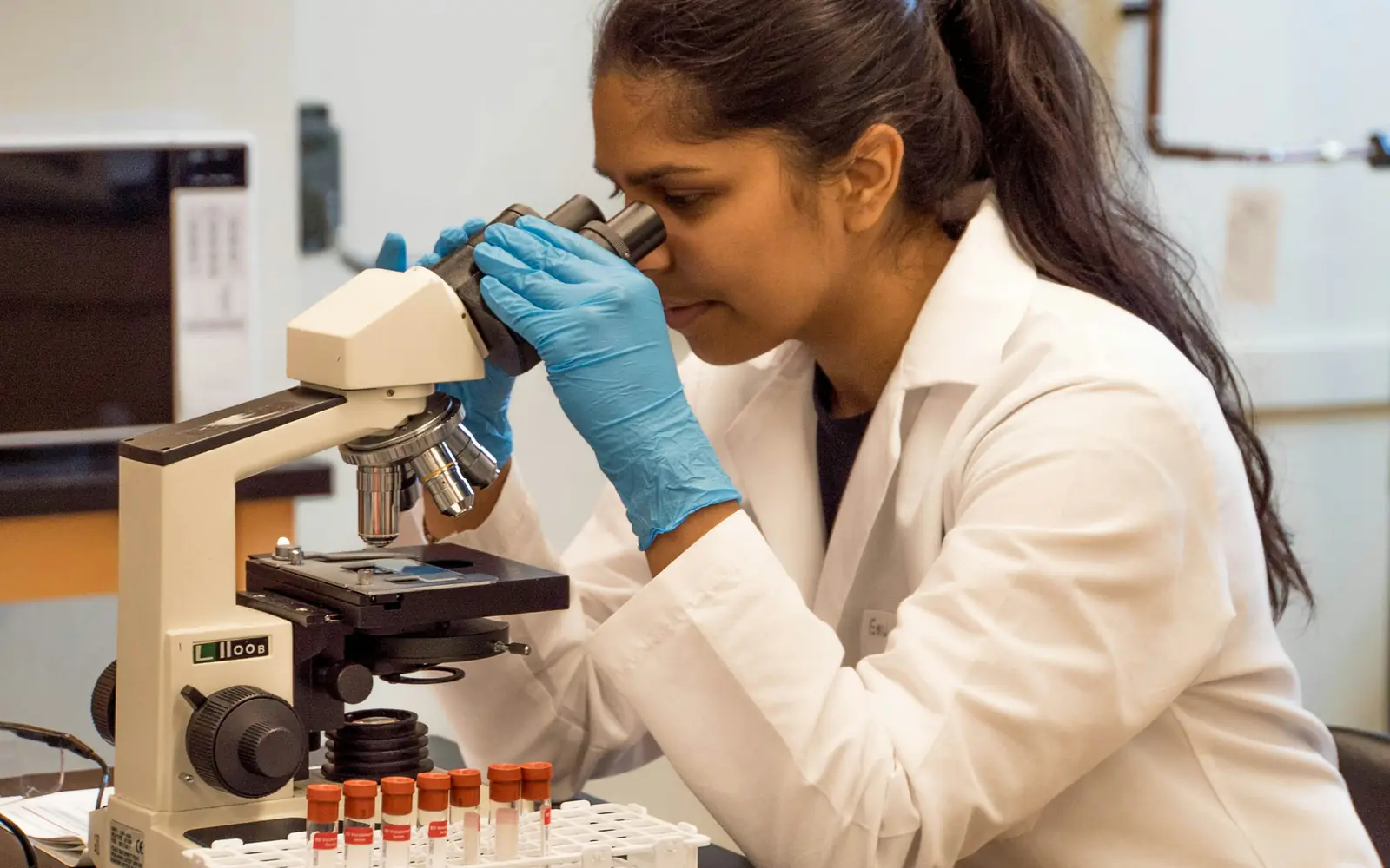 A scientist in a lab coat and blue gloves looks through a microscope beside a rack of sample tubes.