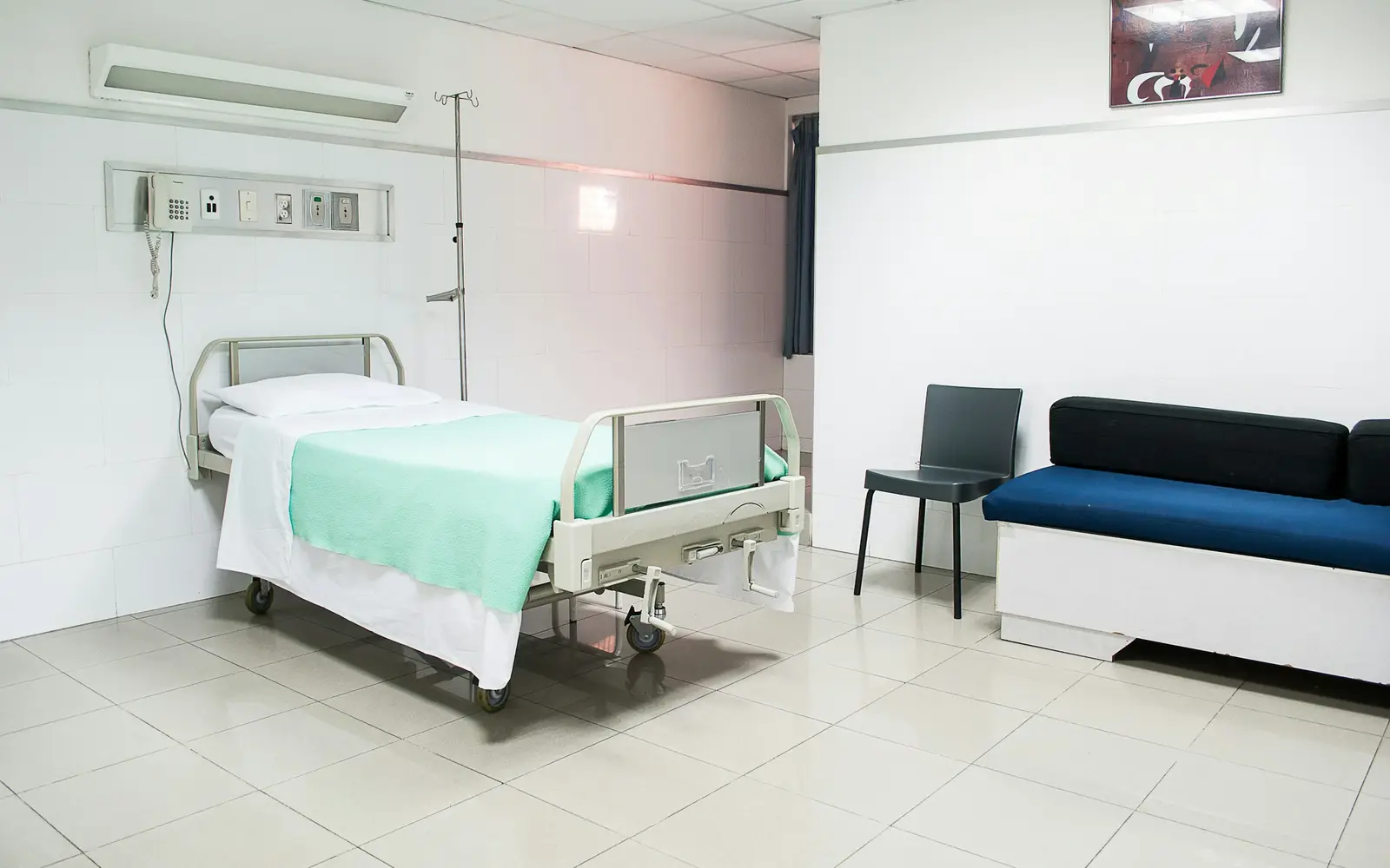 A clean single hospital room with a bed made up with a green blanket and a navy sofa for a visitor.