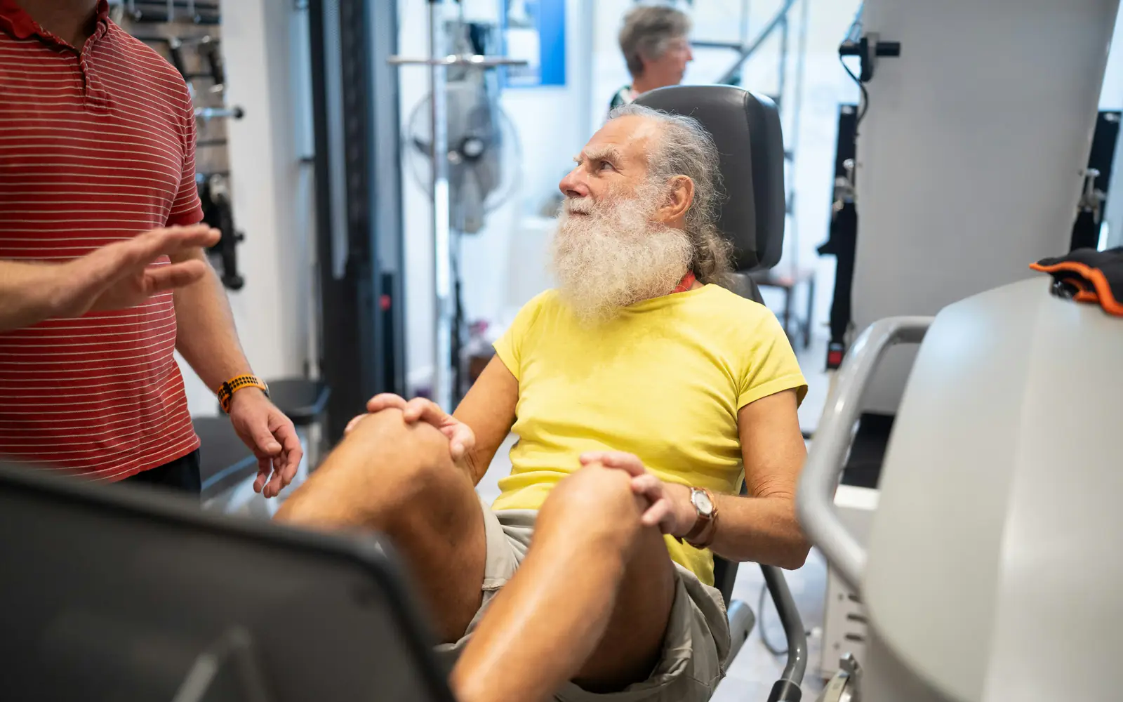 An older man with a white beard sits on an exercise machine with his hands on his knees, talking to a trainer.