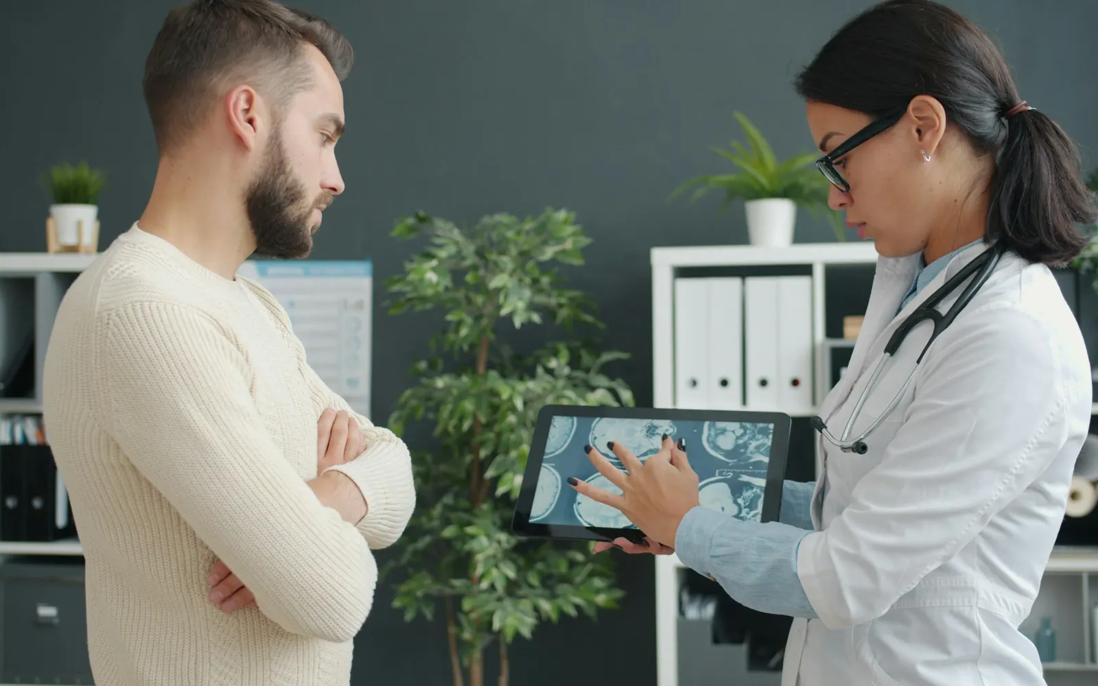 A doctor in glasses and a white coat shows a man scan images on a tablet in a consulting room.