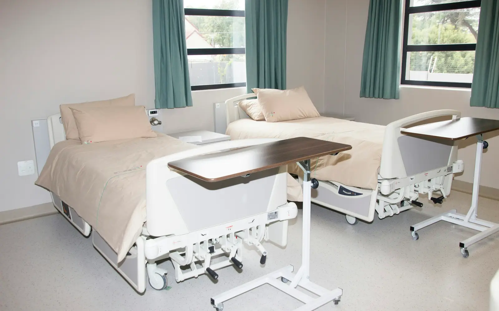 Two made-up hospital beds with over-bed tables in a bright room with teal curtains.