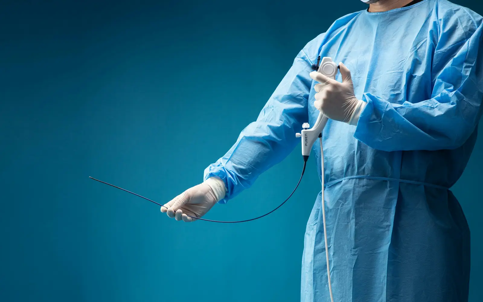 A gloved hand holding a thin, flexible endoscope against a teal background.
