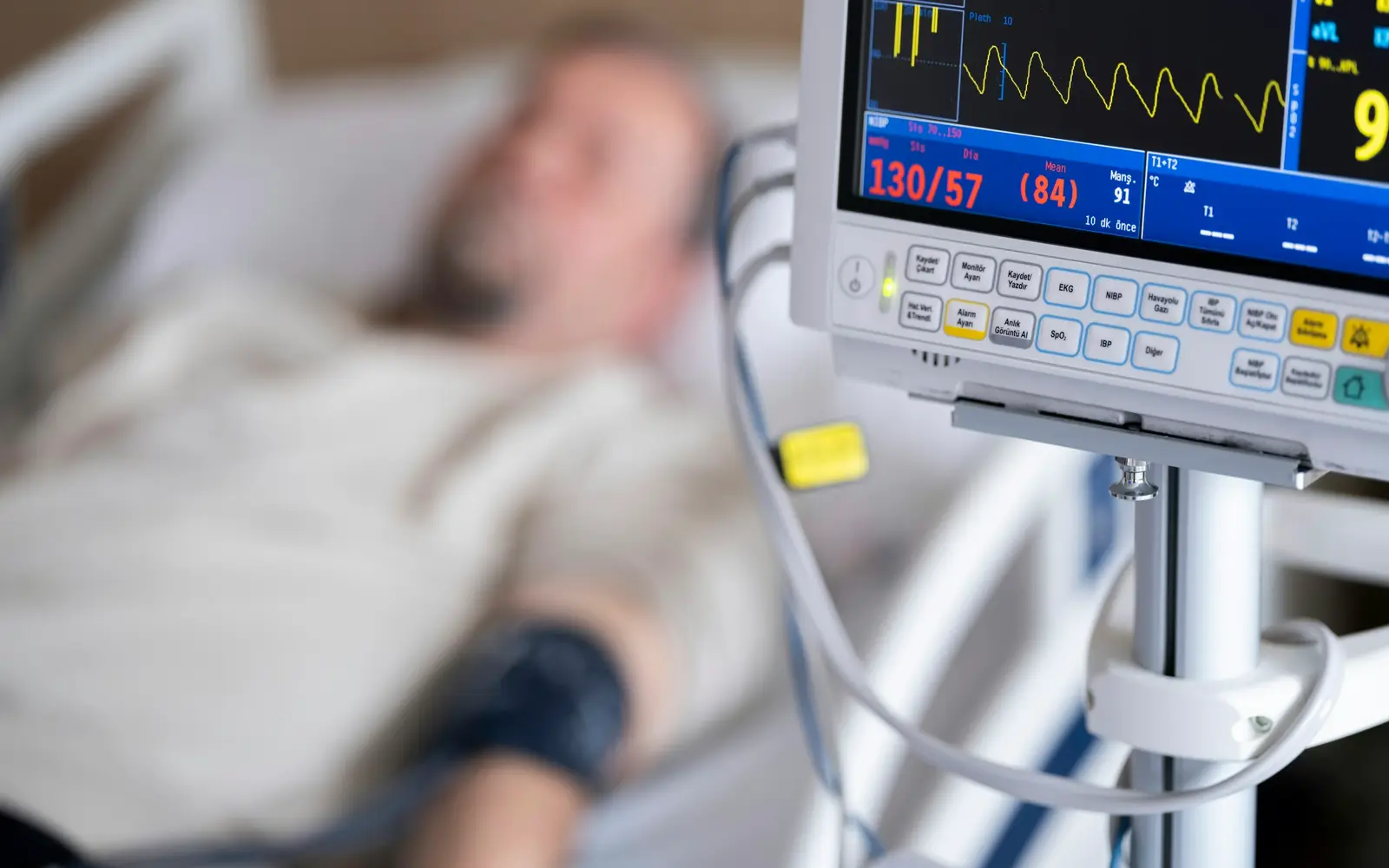 A patient resting in a hospital bed, out of focus, beside a heart monitor showing an ECG trace and blood pressure.