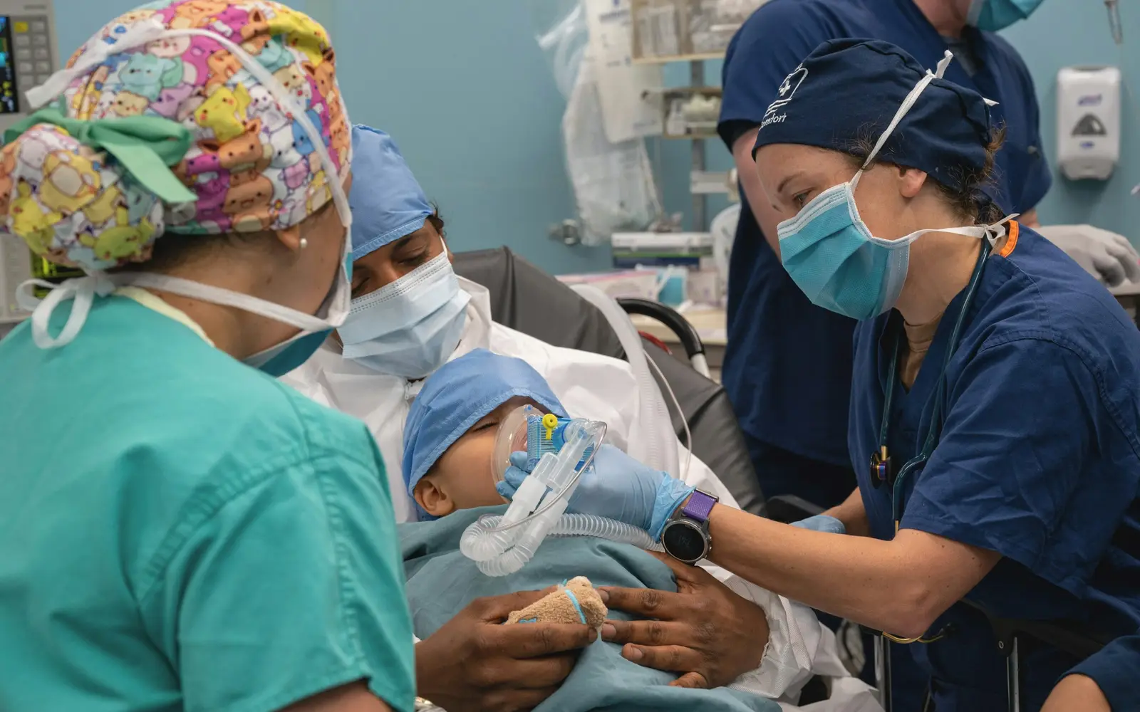 A medical team in scrubs and masks gently holds an oxygen mask for a baby wrapped in a blanket.
