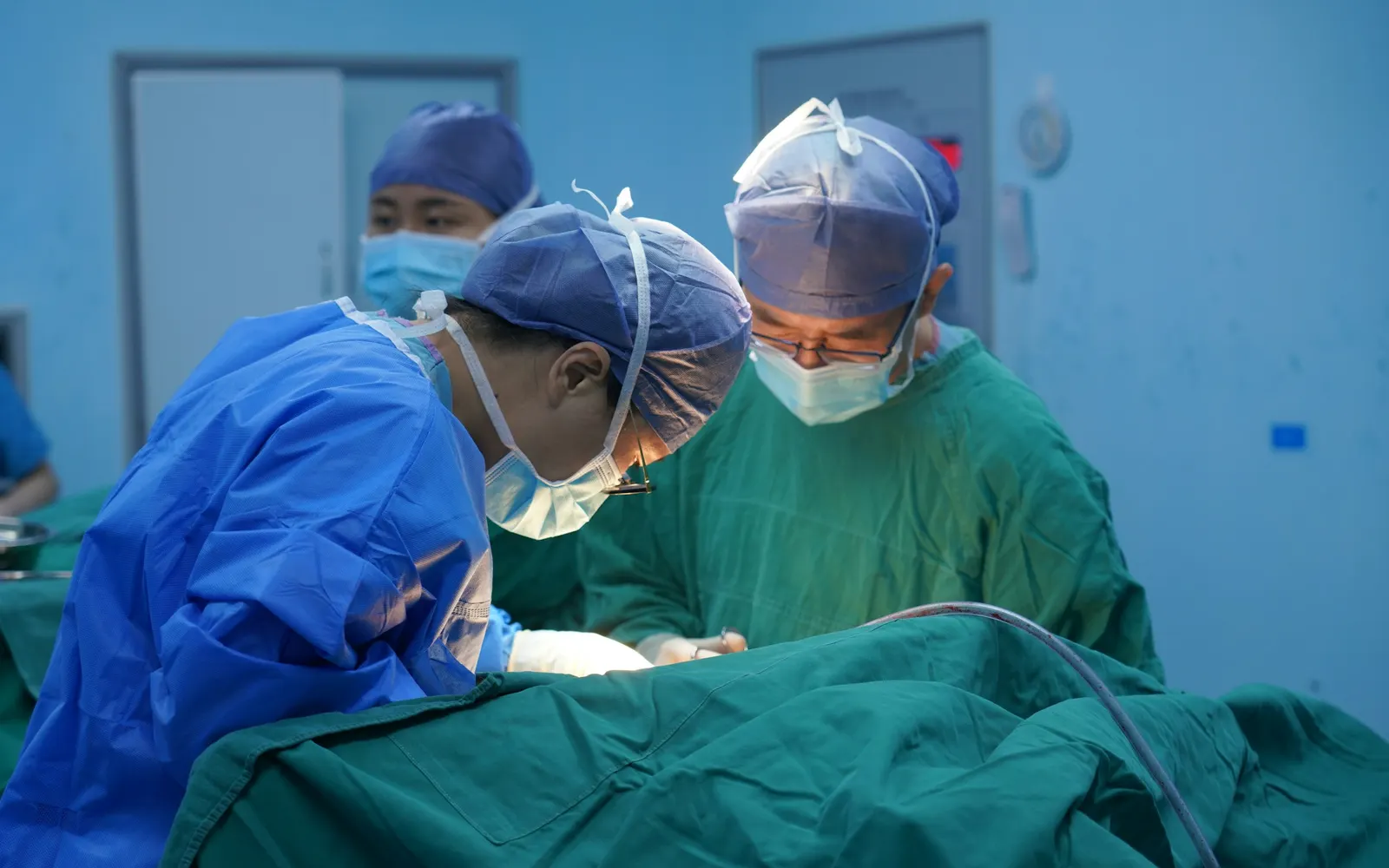 Three surgeons in caps and masks operate on a patient covered with green drapes in an operating theatre.