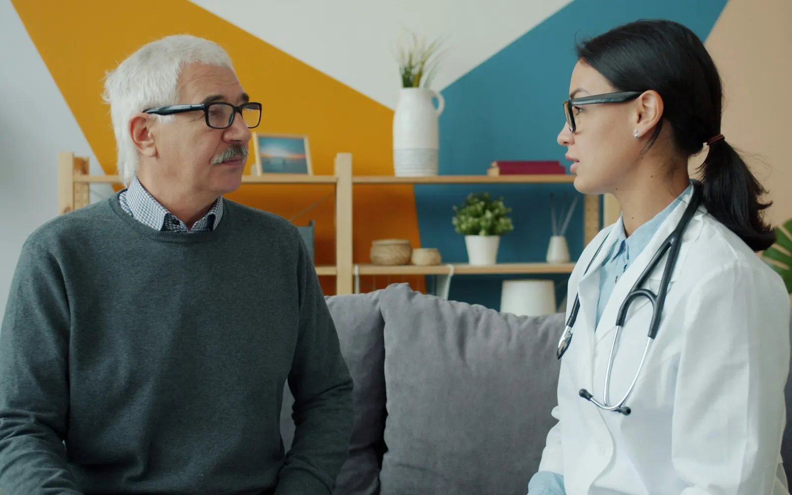 A female doctor in a white coat talks with an older man in a grey jumper as they sit on a sofa.