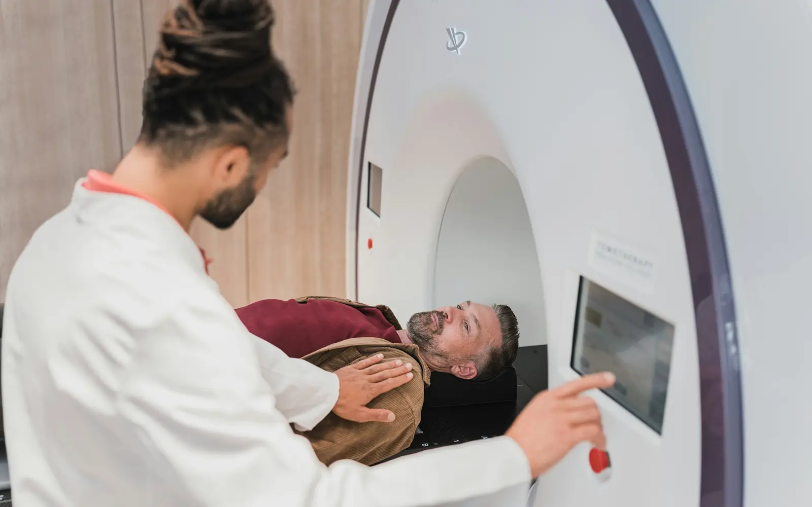 A clinician in a white coat positions a man on the couch of a radiotherapy machine and uses its touchscreen.