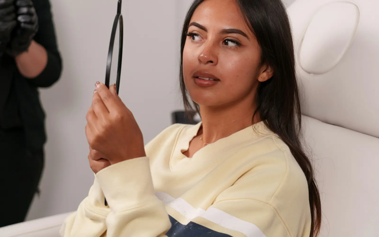 A young woman in a clinic chair looks at her face in a hand mirror.