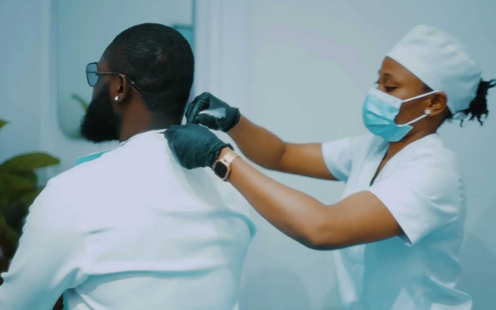 A nurse in a mask and gloves examines the upper back and shoulders of a seated patient.