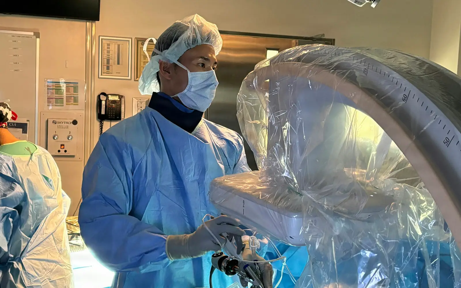 A surgeon in a mask and cap works beside draped imaging equipment in an operating room.