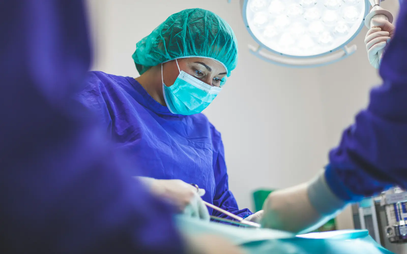 A surgeon in blue scrubs, a green cap and a mask operates under a bright theatre light.